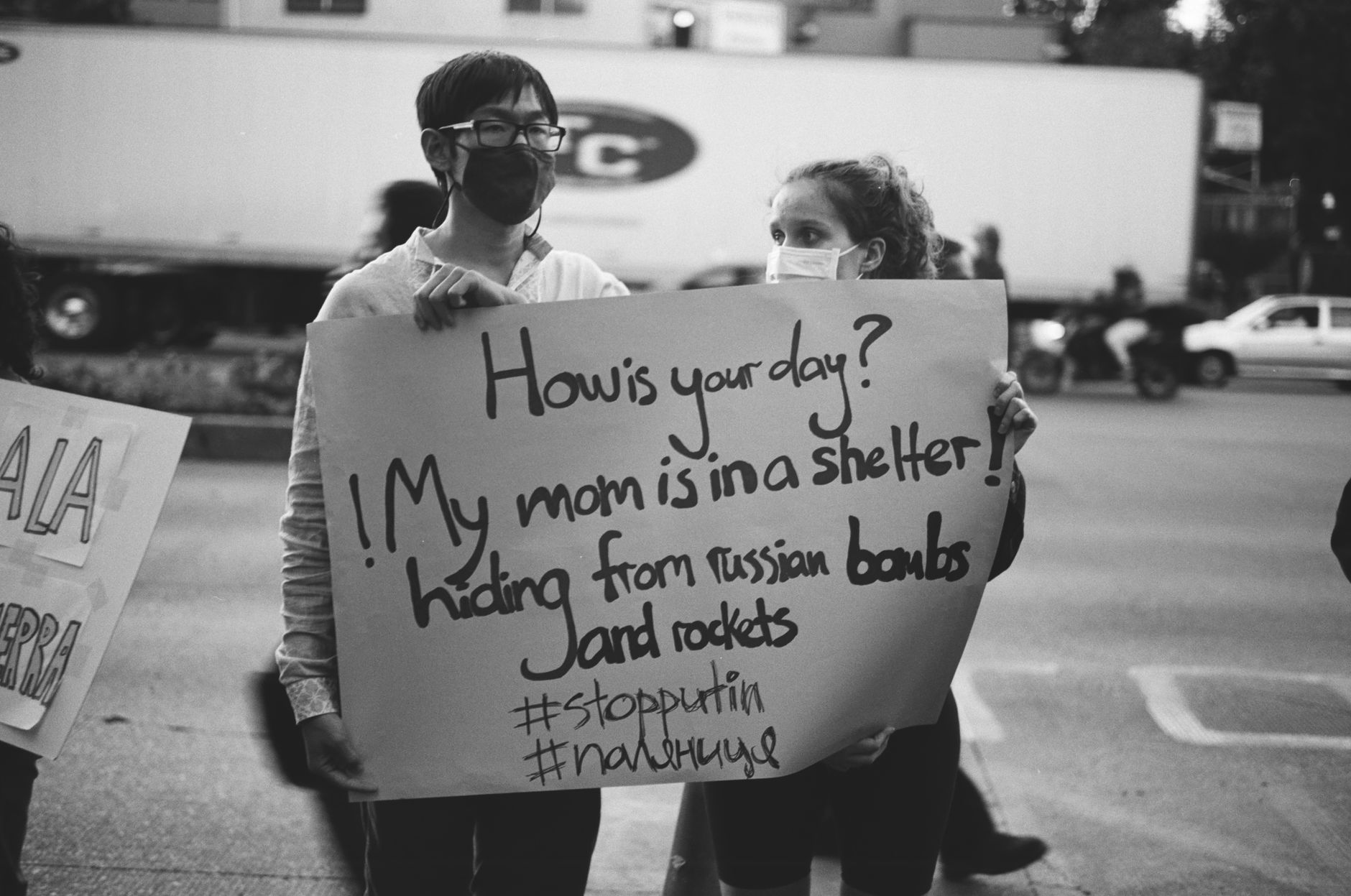 Two masked individuals hold a protest sign about shelter concerns amidst a street scene.